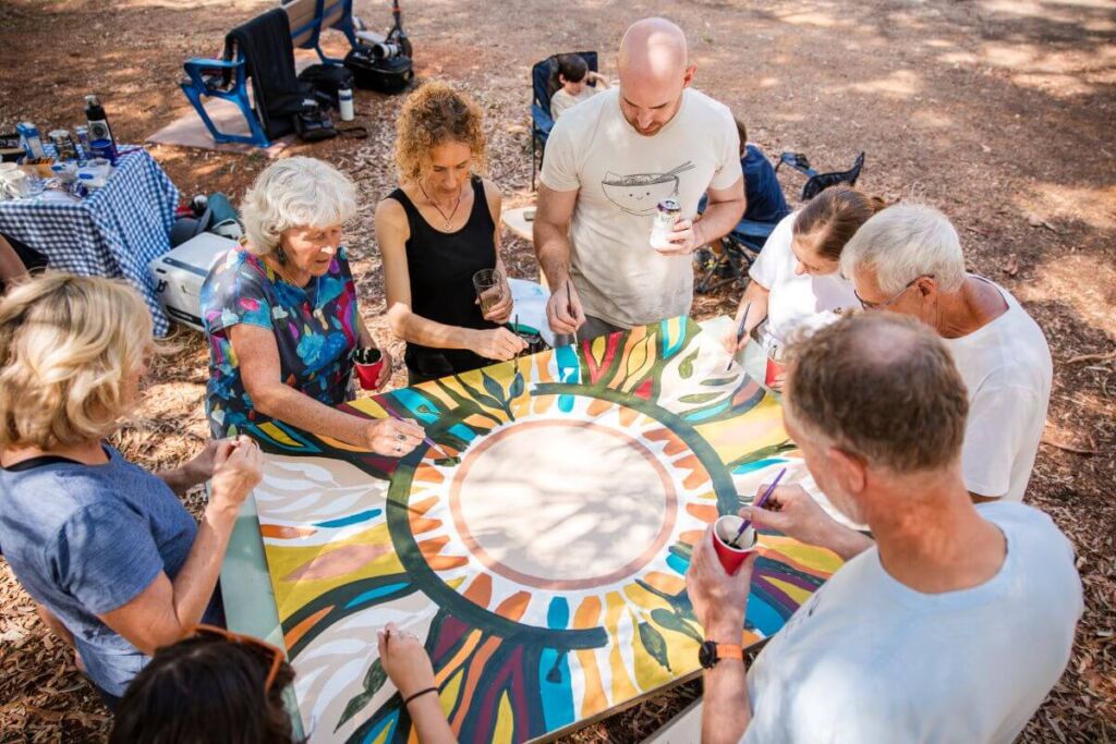A group of people of mixed ages collaboratively painting a large, colorful mandala on a wooden board during an outdoor art workshop in a forest setting.