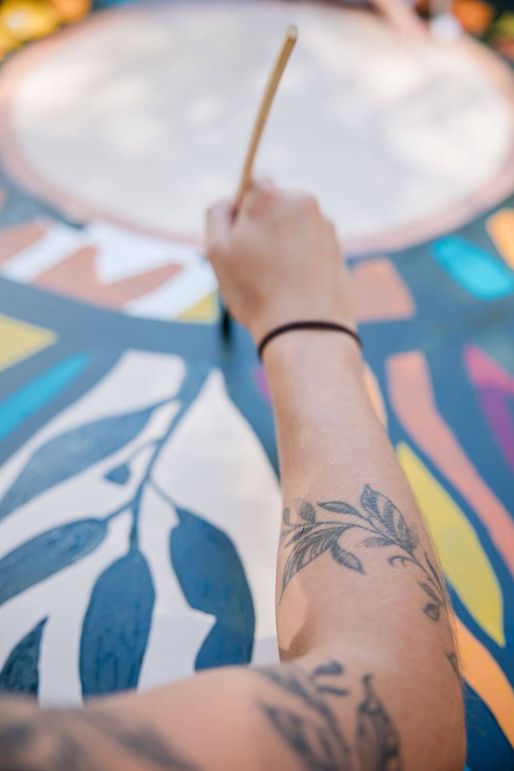 Tattooed arm painting leafy patterns on a colourful circular mural.